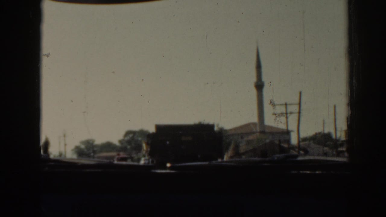 Moving 8mm view in Izmir, Turkey, with a dark truck in the road, a mosque minaret, roadside buildings, and utility poles.