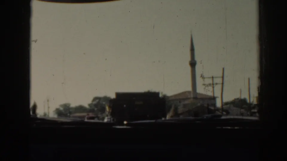 Moving 8mm view in Izmir, Turkey, with a dark truck in the road, a mosque minaret, roadside buildings, and utility poles.