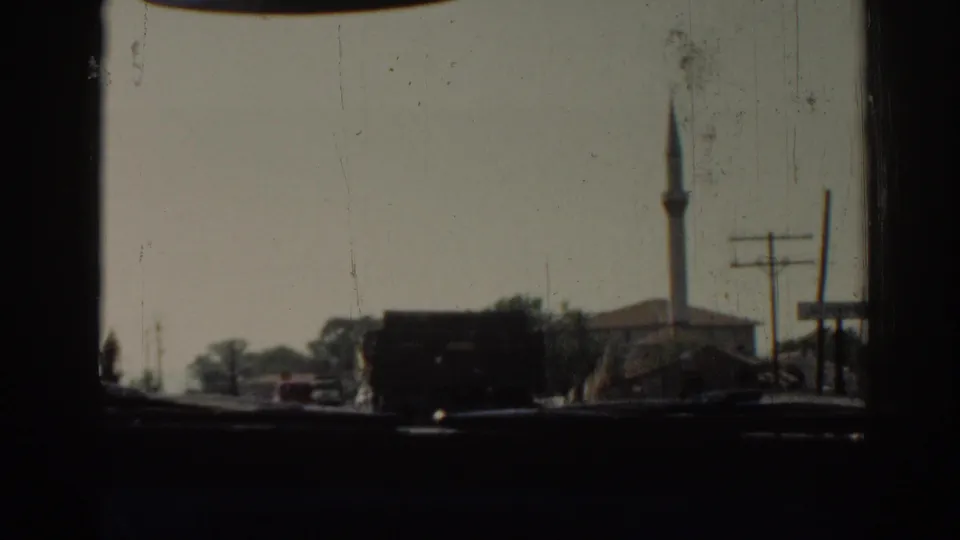 1962 Izmir street scene from inside a car, with a dark truck, small red car, utility poles, trees, and a mosque minaret.