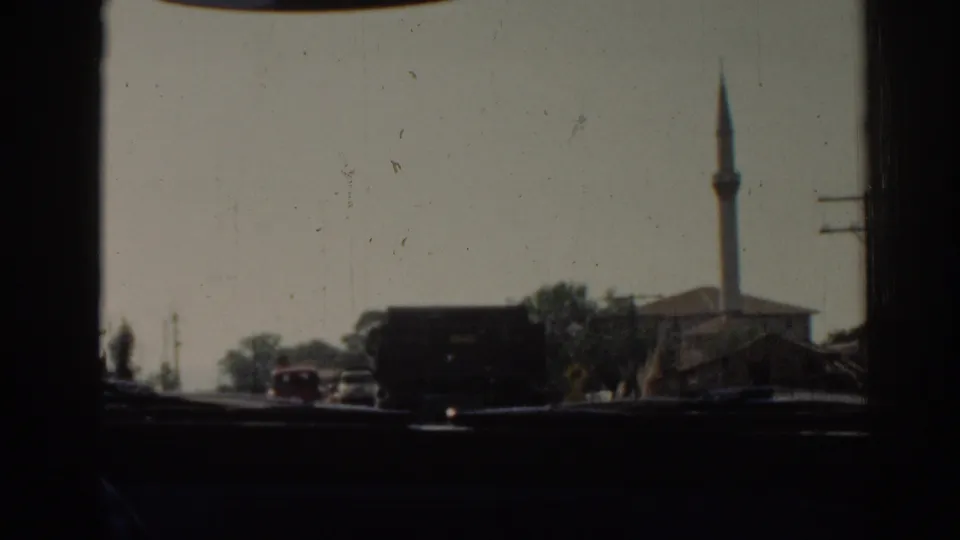 Soft 8mm car-window frame of Izmir street traffic, showing a dark truck, red car, low buildings, trees, and a mosque minaret.