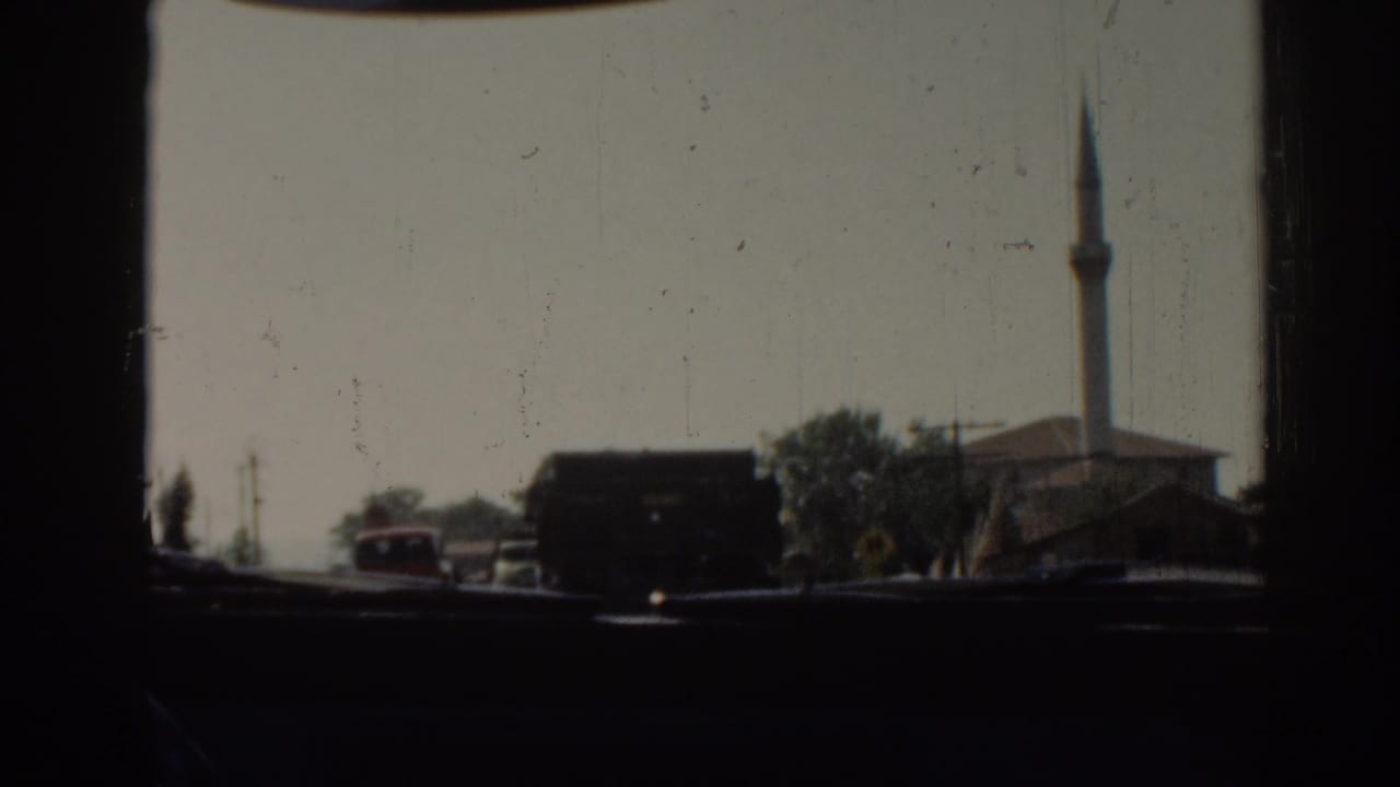 View through a vehicle window of road traffic with trucks, cars, trees, and a distant minaret.