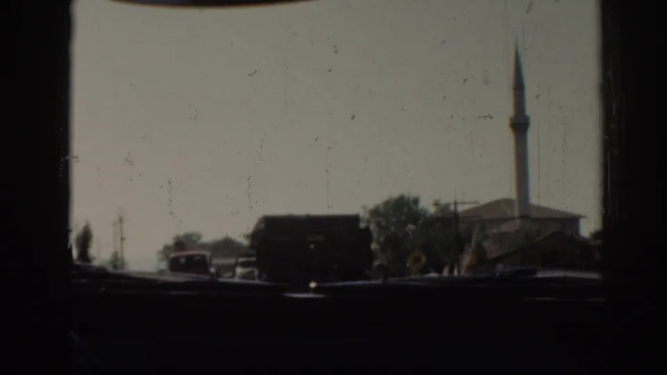 1962 Izmir road scene filmed through a vehicle window, with trucks ahead, roadside trees, and a mosque minaret in the distance.