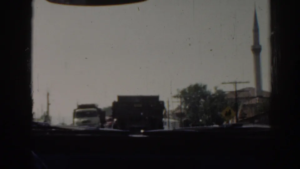 1962 home-movie view from a vehicle in Izmir, showing road traffic, roadside poles, trees, and a tall mosque minaret.