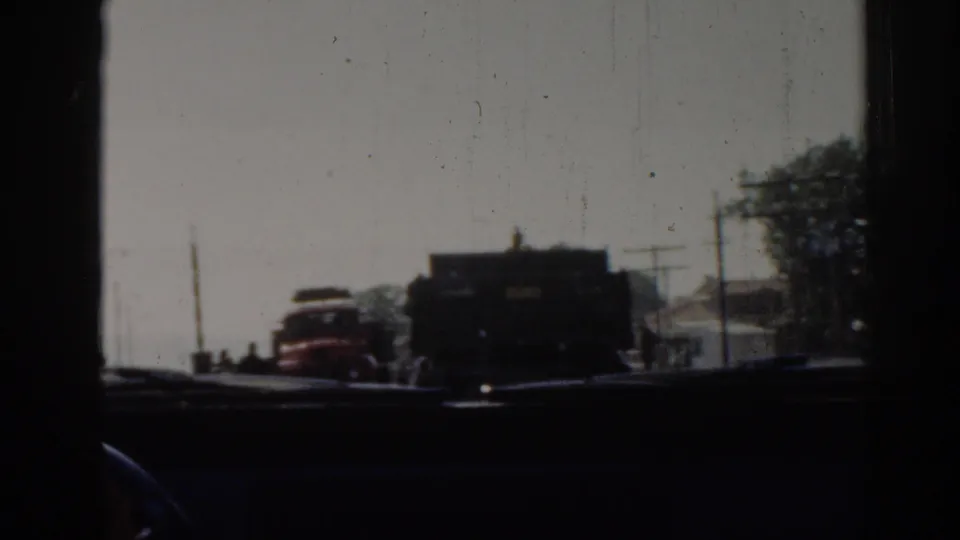 Izmir traffic filmed through a dusty vehicle window in 1962, with a red car, dark truck, utility poles, trees, and a minaret visible.