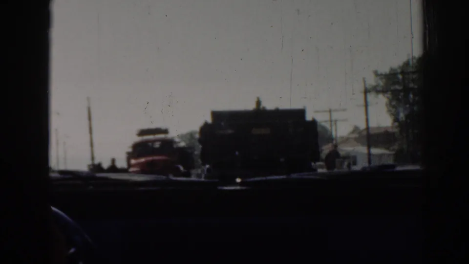 1962 Izmir home-movie frame looking through a windshield at a red truck, a dark truck, utility poles, and roadside buildings.
