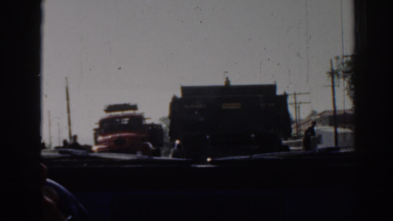 From inside a vehicle in 1962 Izmir, the frame shows a red truck, a dark truck, roadside structures, and small figures near the road.