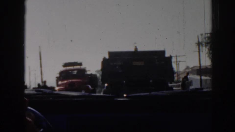 From inside a vehicle in 1962 Izmir, the frame shows a red truck, a dark truck, roadside structures, and small figures near the road.
