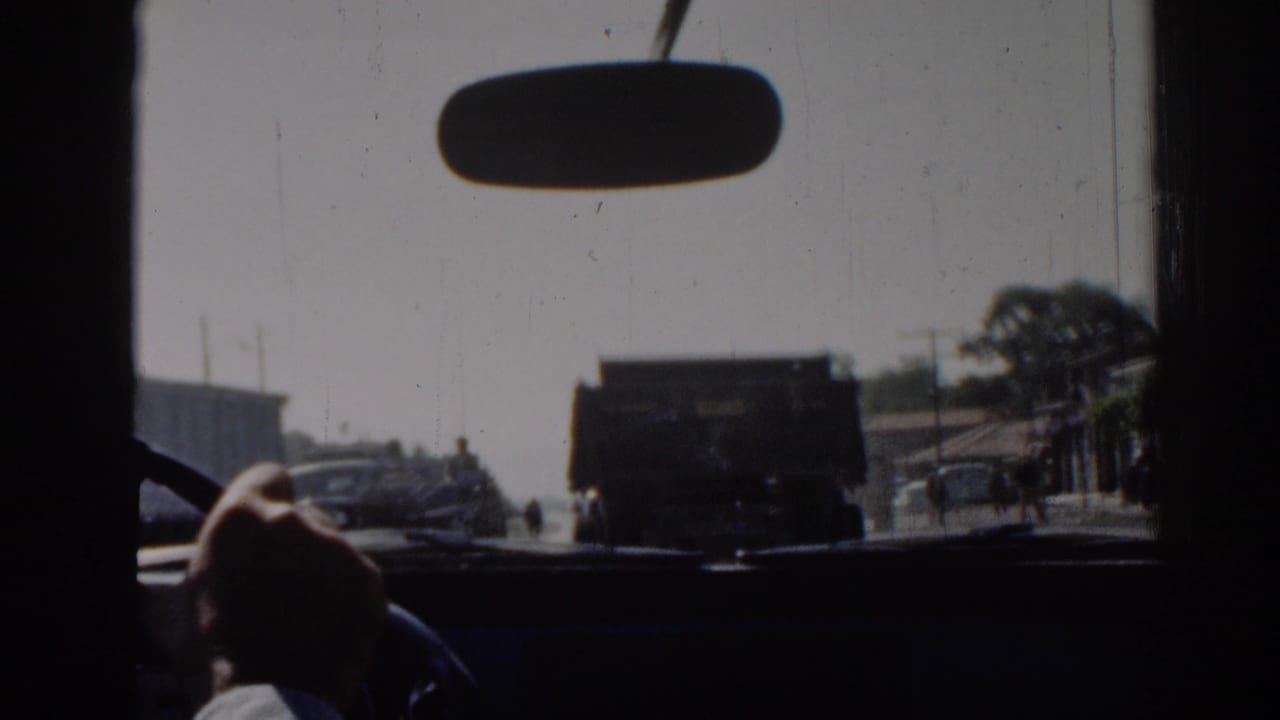 Interior car view in Izmir, 1962, with a rearview mirror, driver's hand on the wheel, and a dark truck ahead on the sunlit road.