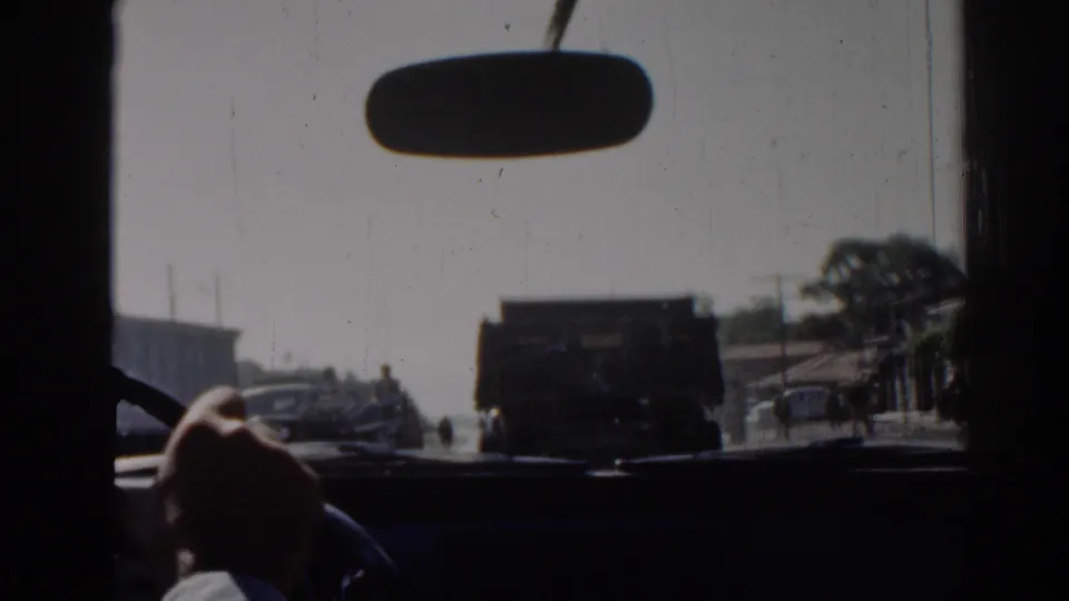 Interior car view in Izmir, 1962, with a rearview mirror, driver's hand on the wheel, and a dark truck ahead on the sunlit road.