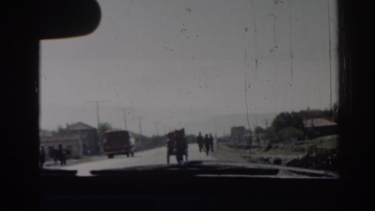 View through a car windshield toward a road with a red van, pedestrians, houses, and utility poles.