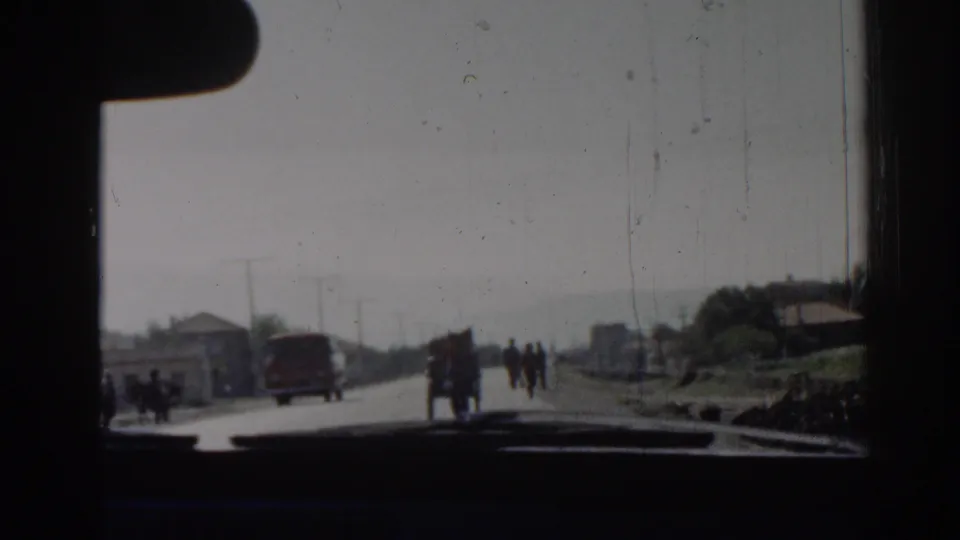 1962 Izmir street seen from inside a moving car, with a red van ahead and pedestrians walking near roadside houses.