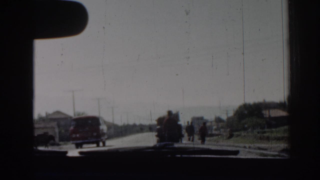 The car approaches a red van on a 1962 Izmir road as small pedestrian groups stand or walk near low roadside buildings.