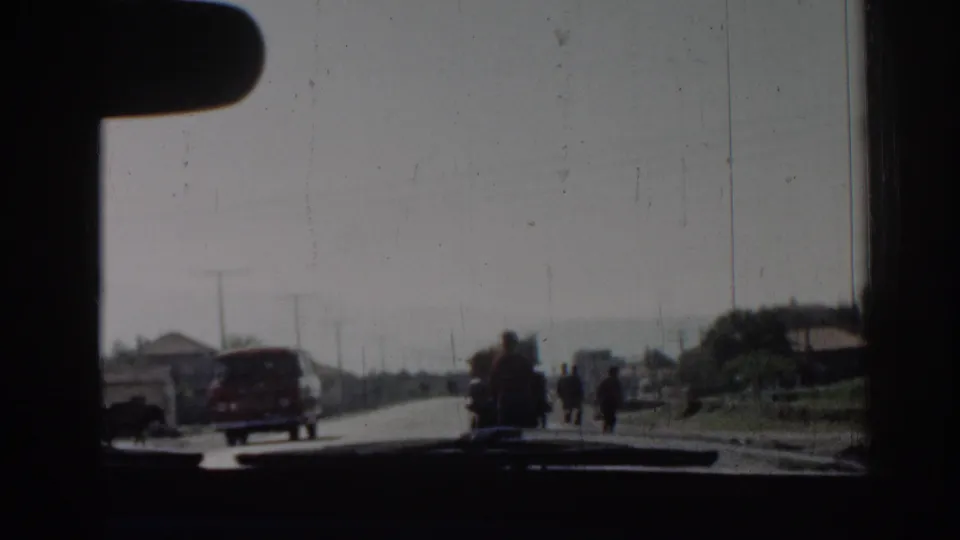 The car approaches a red van on a 1962 Izmir road as small pedestrian groups stand or walk near low roadside buildings.