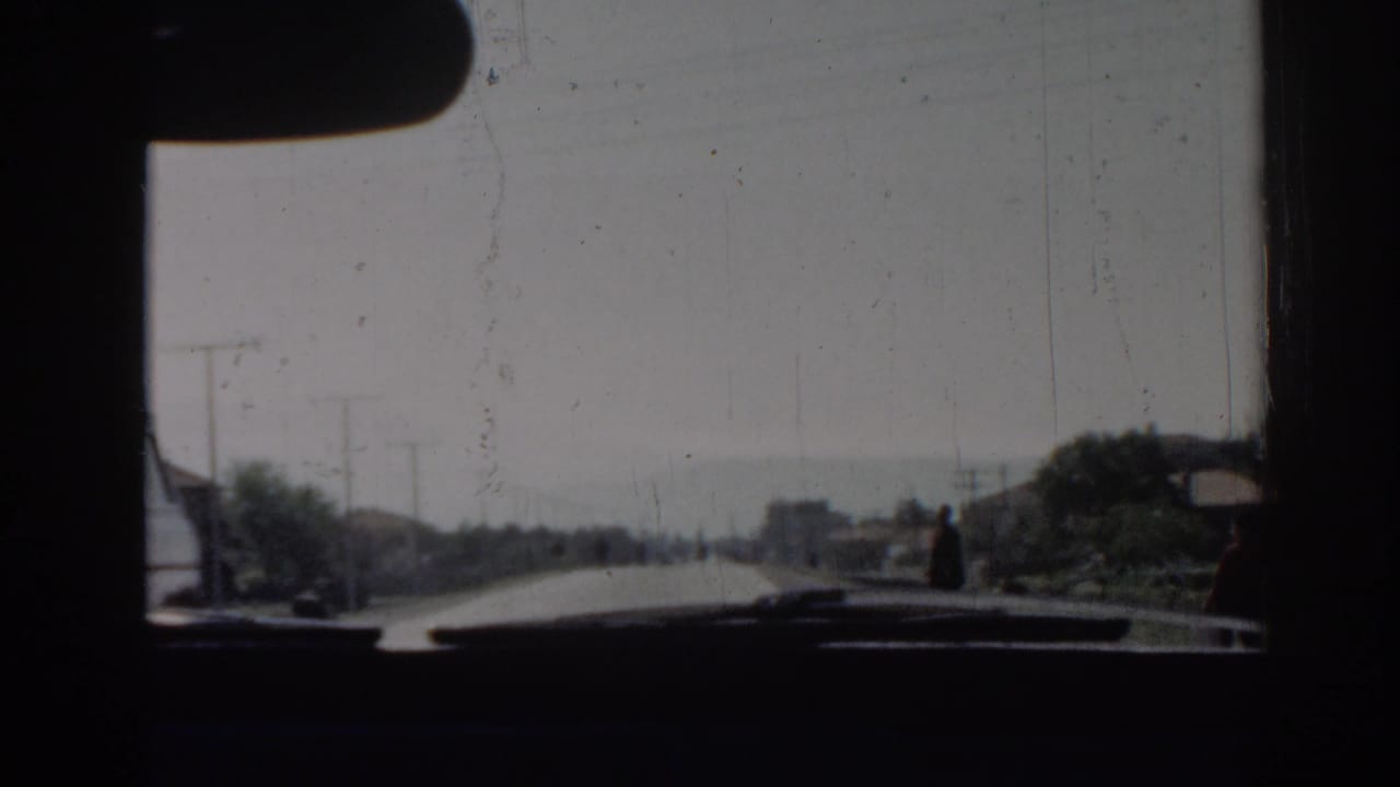 A forward windshield view in 1962 Izmir shows the road opening ahead, with distant pedestrians, roadside houses, and utility poles.