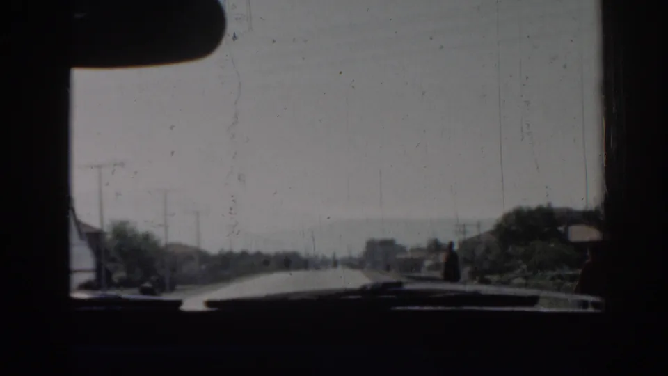 A forward windshield view in 1962 Izmir shows the road opening ahead, with distant pedestrians, roadside houses, and utility poles.