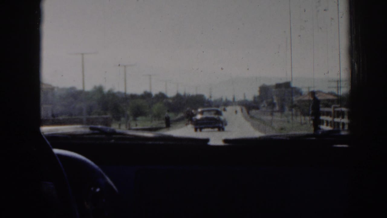 1962 Izmir road scene from inside a vehicle, showing a car ahead, roadside structures, trees, and the dark dashboard foreground.