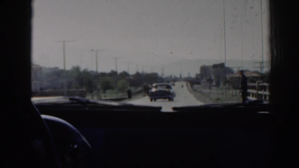 1962 Izmir road scene from inside a vehicle, showing a car ahead, roadside structures, trees, and the dark dashboard foreground.