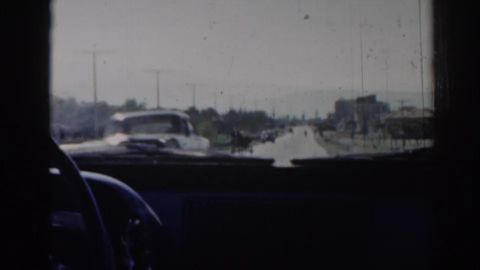 From inside a car in Izmir, Turkey, the 1962 footage shows an oncoming vehicle, distant road users, poles, trees, and dashboard shadows.