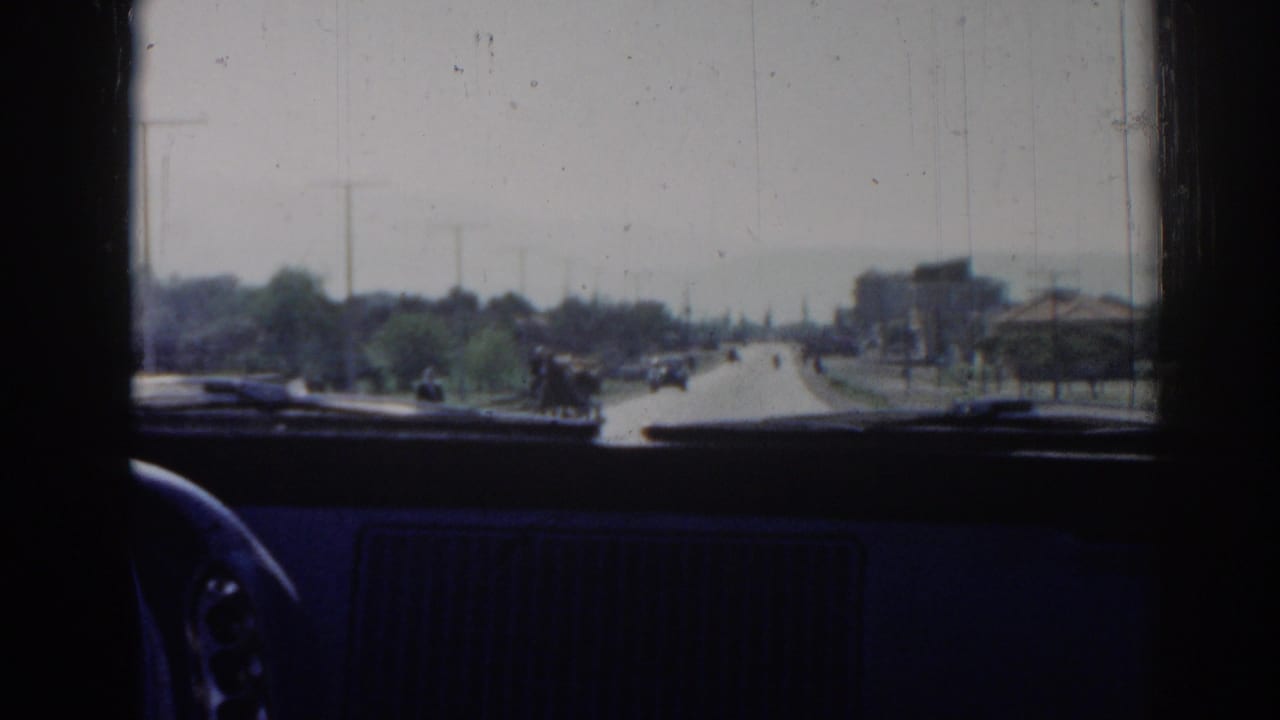 1962 Izmir windshield view showing a sunlit roadway, distant pedestrians or riders, scattered cars, trees, and low roadside buildings.
