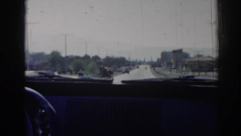 1962 Izmir windshield view showing a sunlit roadway, distant pedestrians or riders, scattered cars, trees, and low roadside buildings.