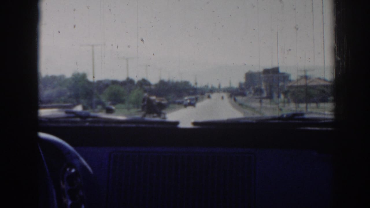 View through a car windshield onto a sunlit road with vehicles, trees, and utility poles.