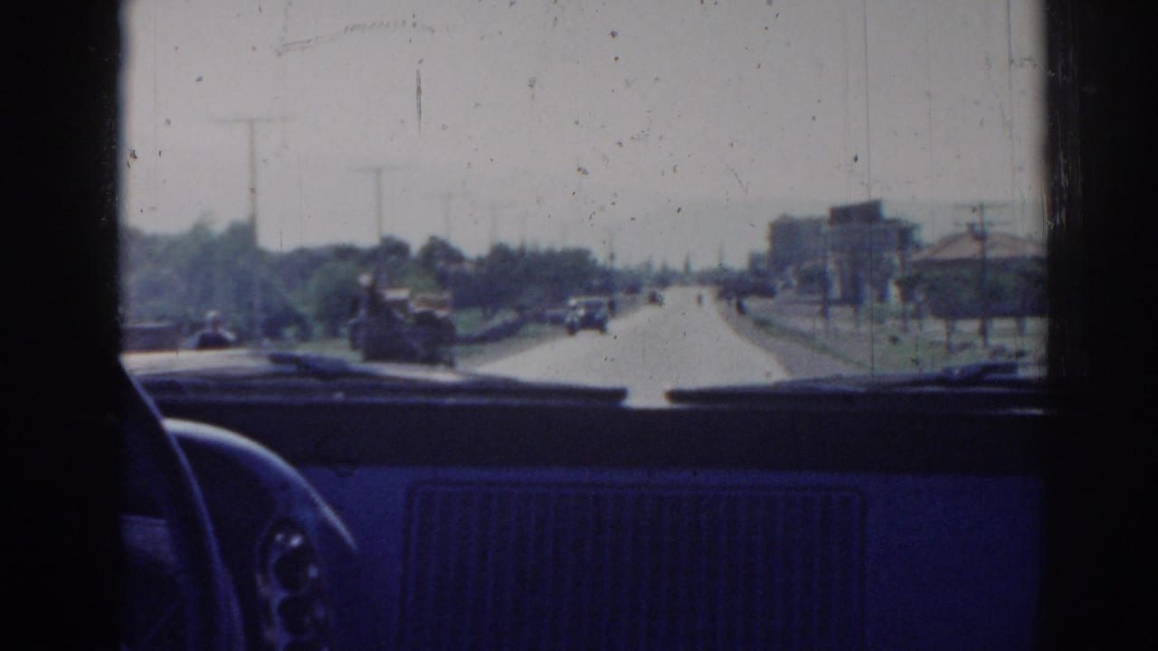 Interior car perspective on a 1962 Izmir roadway, with small vehicles ahead, roadside trees, poles, and distant buildings.