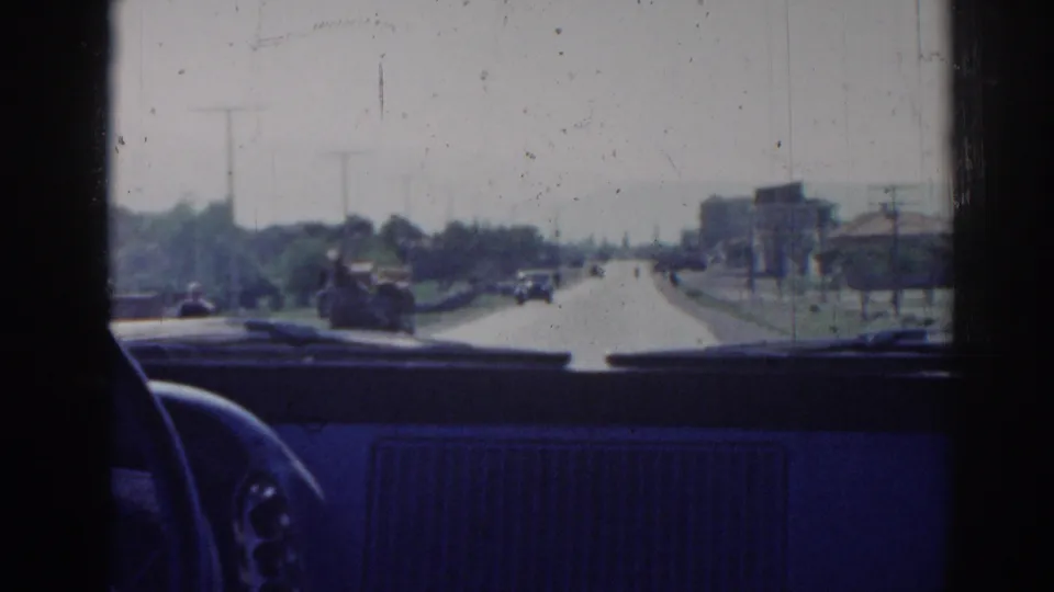 Interior car perspective on a 1962 Izmir roadway, with small vehicles ahead, roadside trees, poles, and distant buildings.