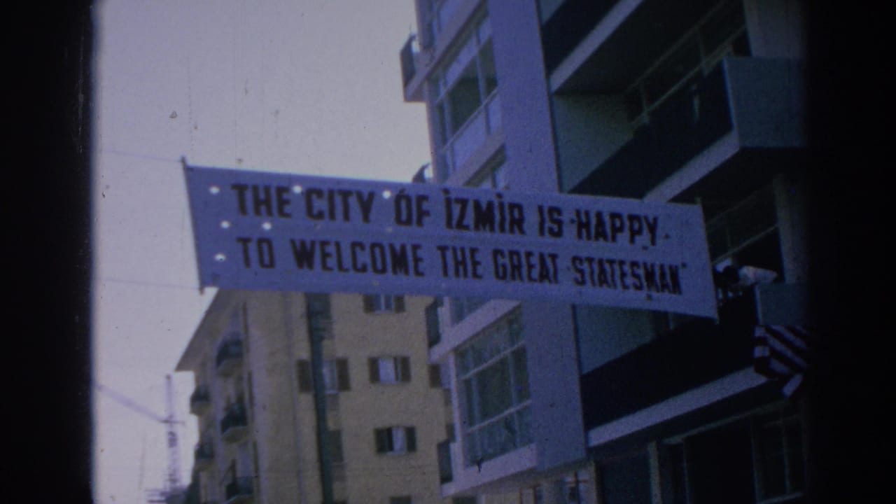 Close 1962 Izmir home-movie frame of a banner reading welcome to the great statesman, with balconies and buildings behind it.