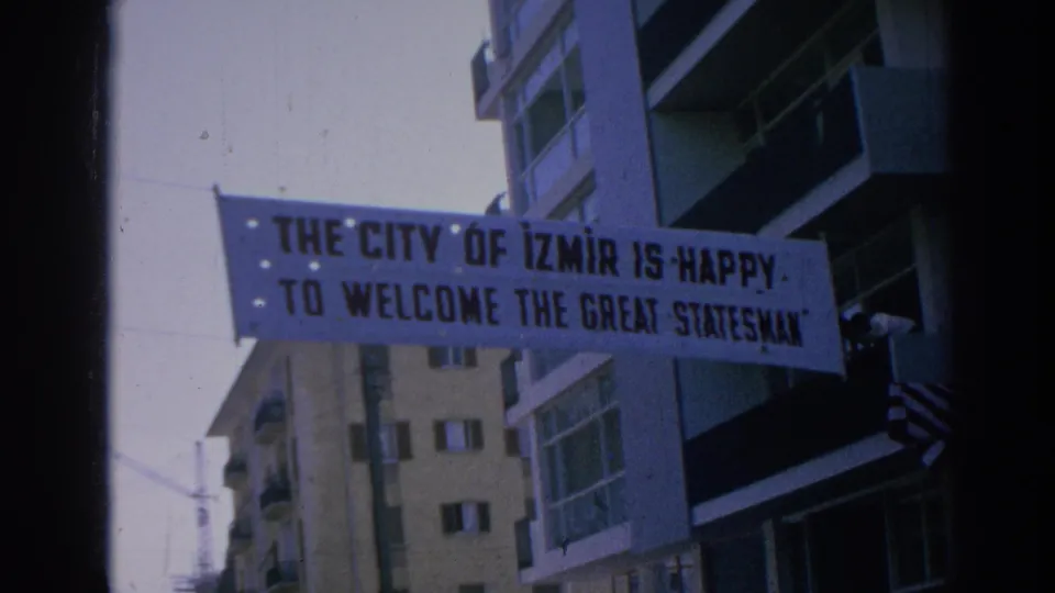 Close 1962 Izmir home-movie frame of a banner reading welcome to the great statesman, with balconies and buildings behind it.