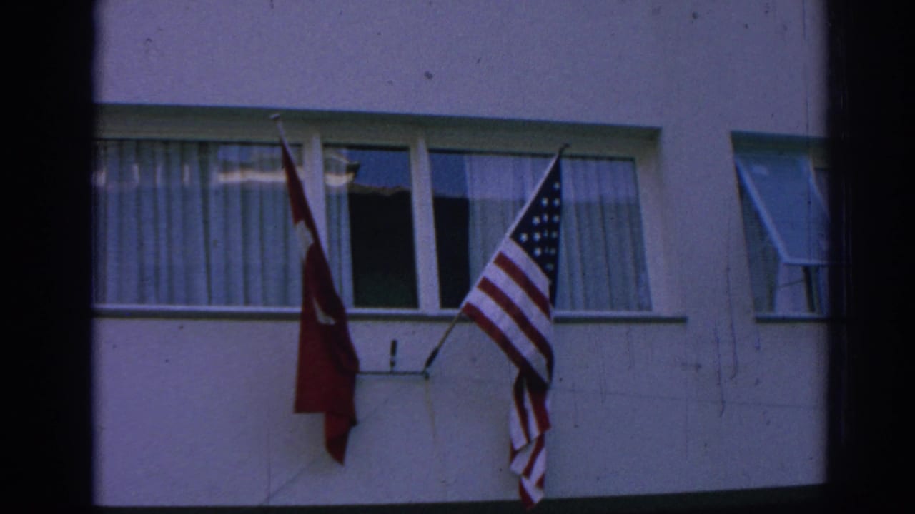 Close home-movie view of Turkish and American flags mounted below a curtained window on a pale building facade.