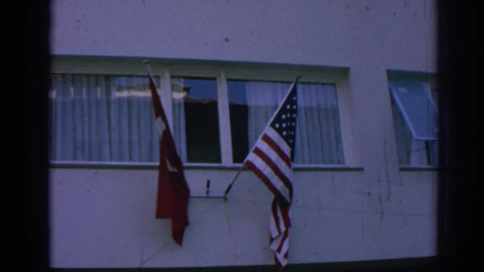 Close home-movie view of Turkish and American flags mounted below a curtained window on a pale building facade.