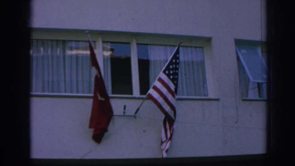 1962 Izmir home-movie frame showing Turkish and American flags fixed outside curtained windows on a light-colored building.