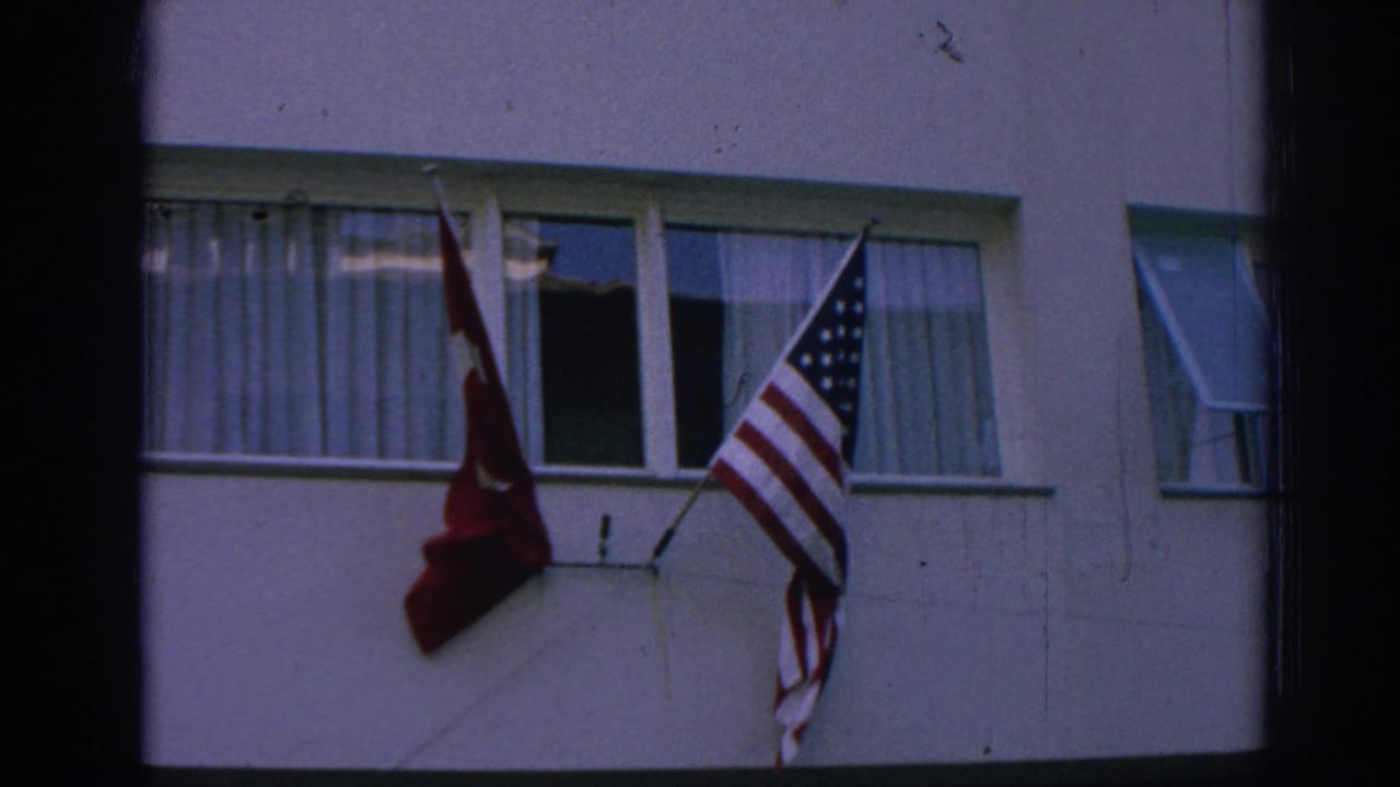 1962 Izmir frame with Turkish and American flags waving from wall brackets beside curtained and open windows.