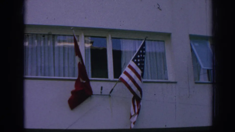 1962 Izmir frame with Turkish and American flags waving from wall brackets beside curtained and open windows.
