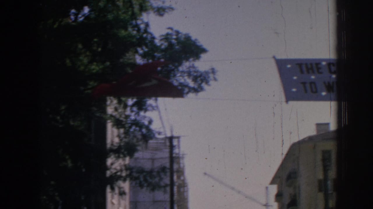 1962 Izmir street-level view with a Turkish flag, trees, urban buildings, and part of a civic welcome banner.