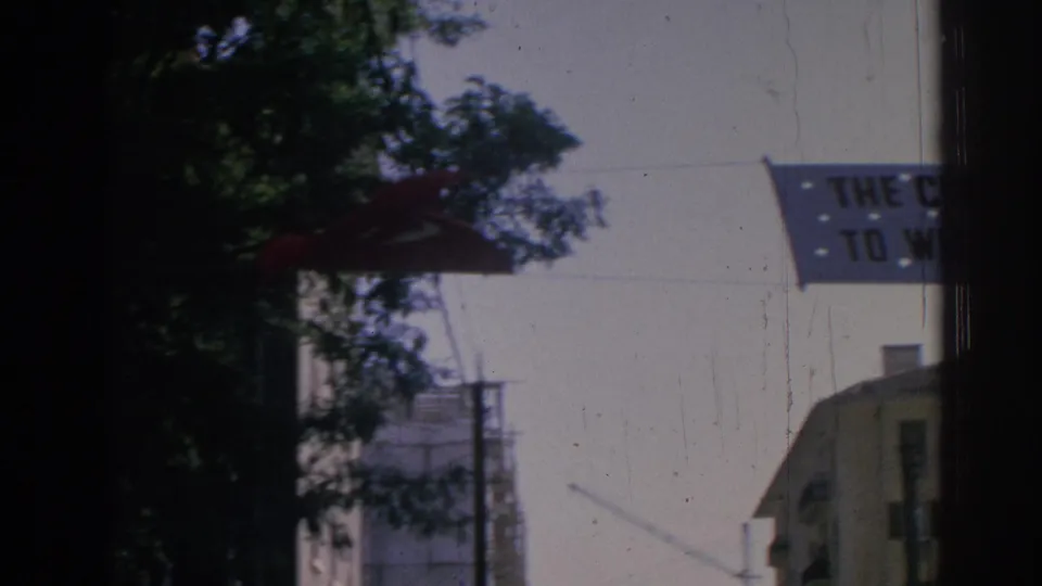 1962 Izmir street-level view with a Turkish flag, trees, urban buildings, and part of a civic welcome banner.