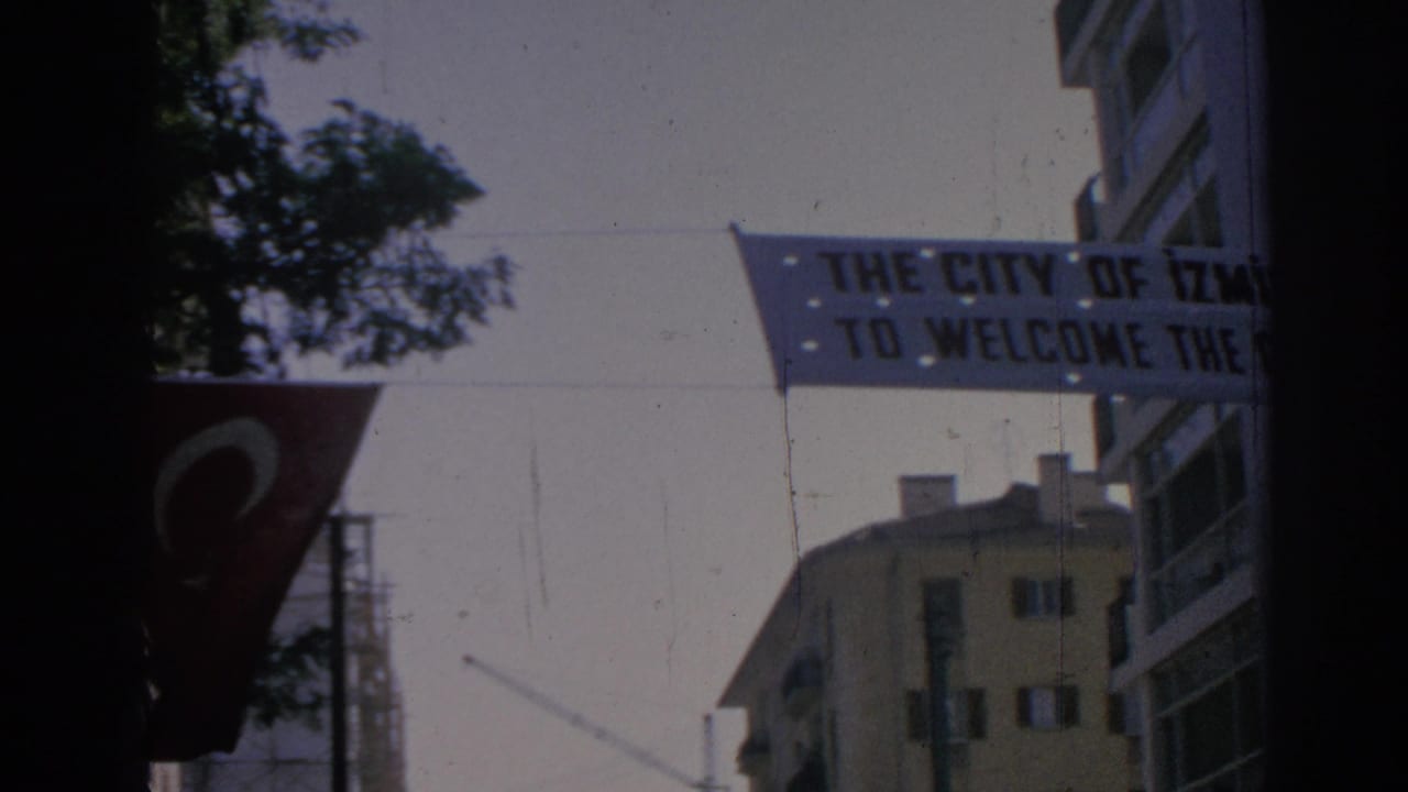 1962 Izmir frame showing a civic welcome banner over a street, with a Turkish flag, trees, and nearby buildings.
