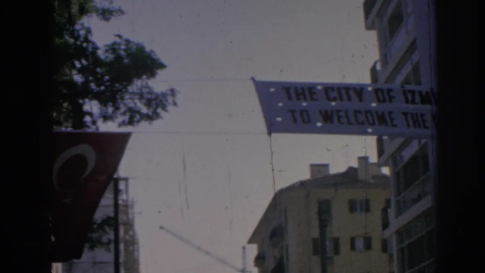 1962 Izmir frame showing a civic welcome banner over a street, with a Turkish flag, trees, and nearby buildings.