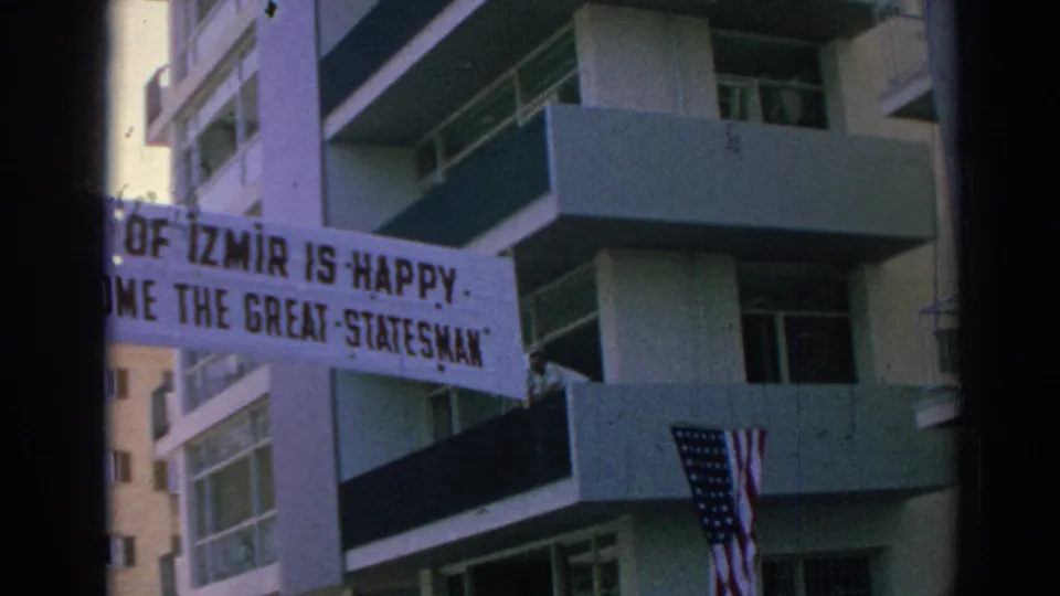 Izmir welcome banner beside apartment balconies, with Turkish and American flags visible below in the 1962 clip.
