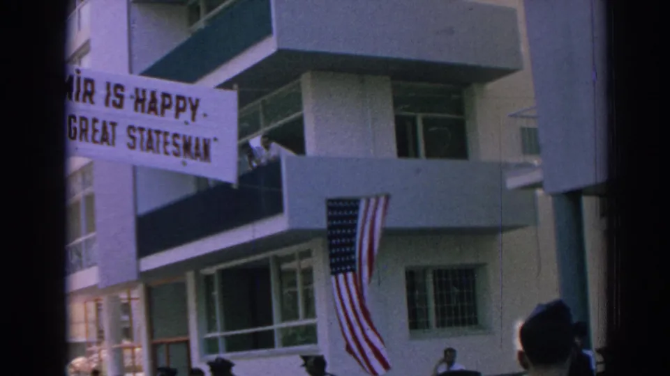 1962 Izmir street view with a welcome banner, American flag, balconies, and figures near the building entrance.