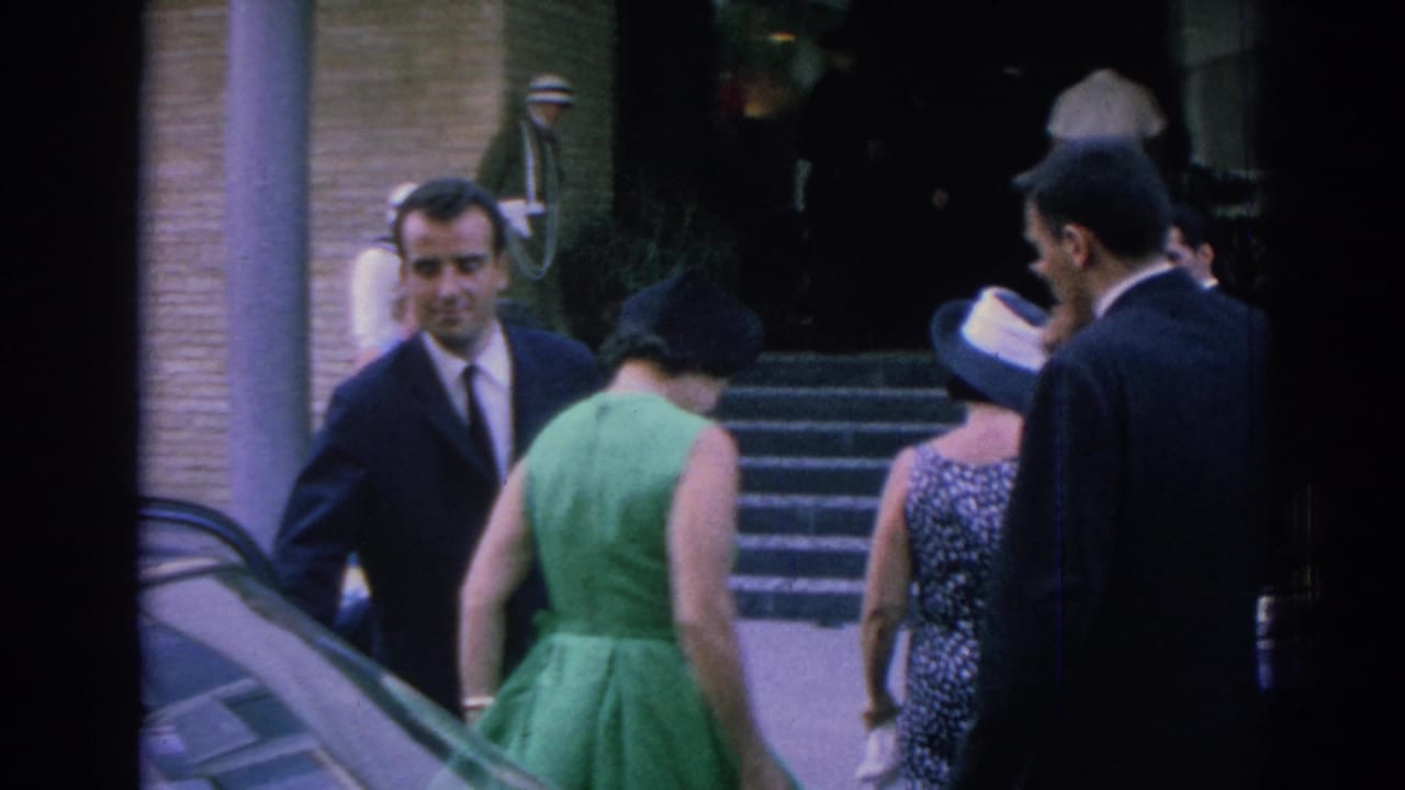 1962 Izmir frame showing a woman in a green dress moving away from a car as suited men and another woman approach the entrance.
