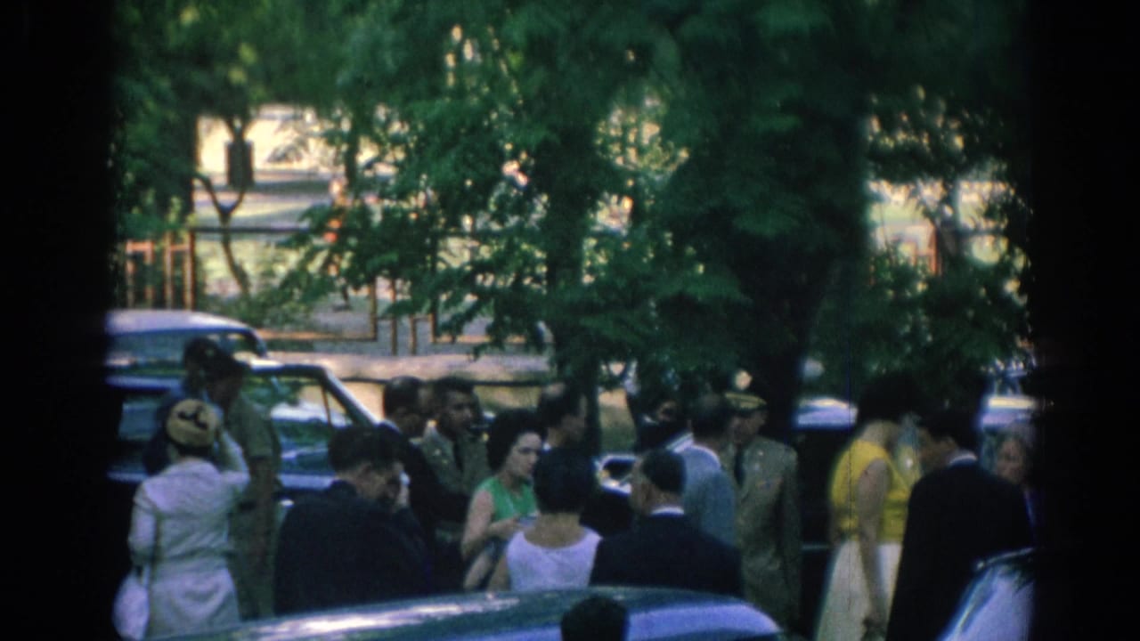 Crowd near trees and a car as uniformed officers and suited men stand nearby.