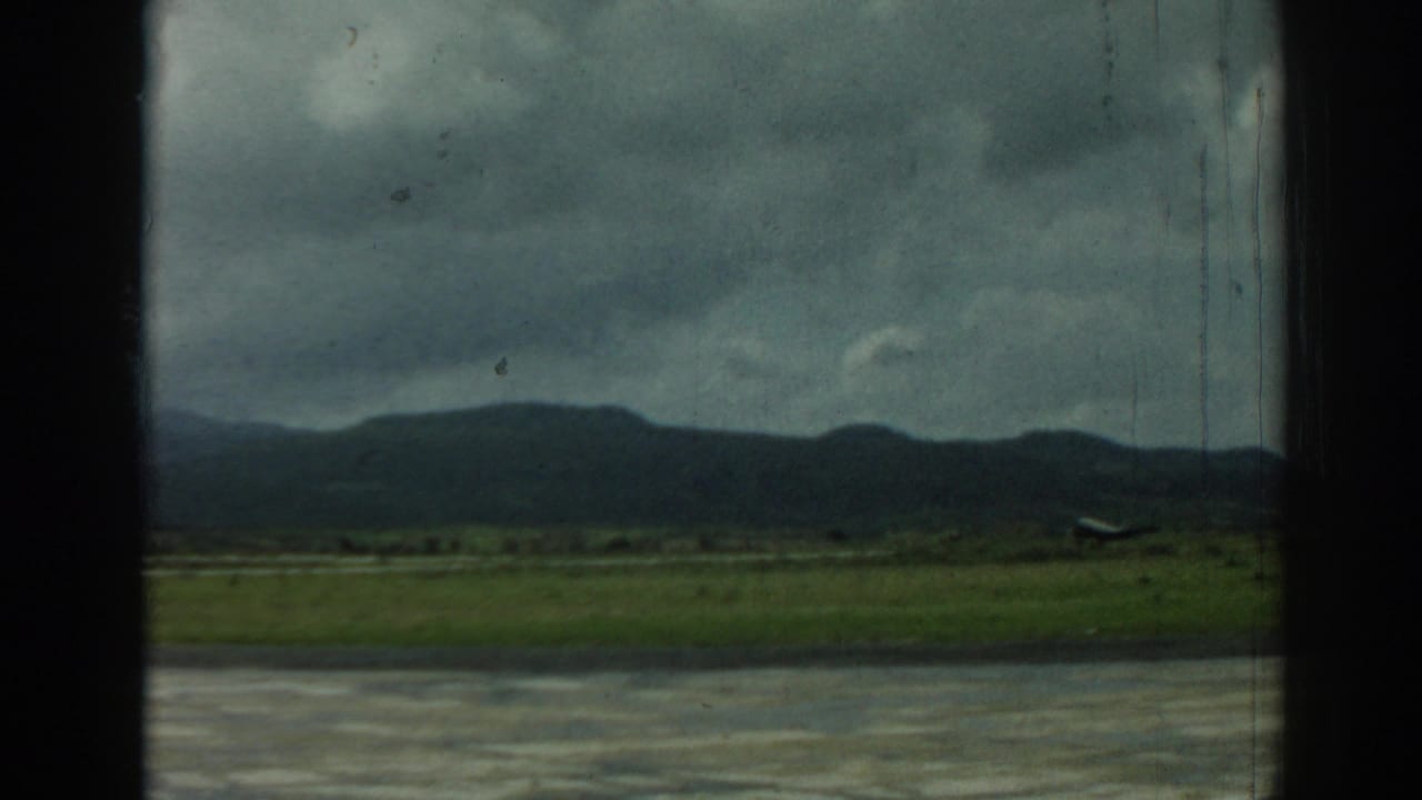 Small aircraft on a runway with grass, mountains, and cloudy sky.