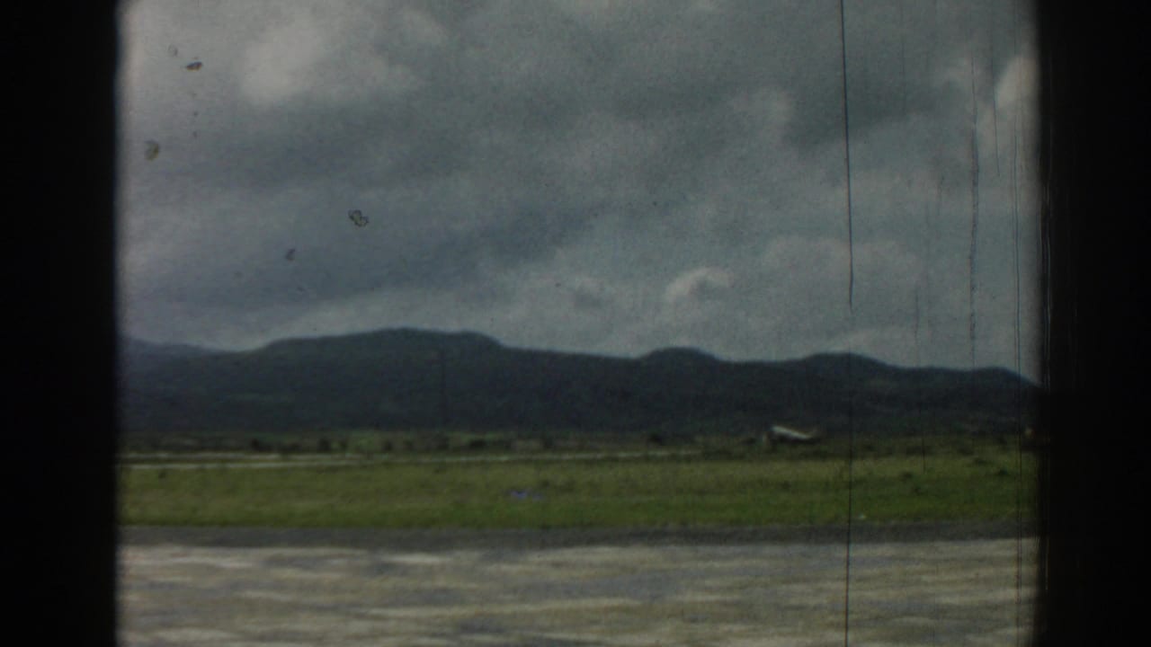 A small aircraft is seen near the runway at the Izmir airfield, framed by grass, distant mountains, overcast sky, and visible film wear.