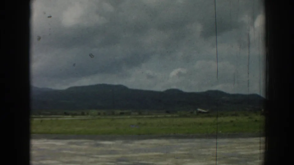 A small aircraft is seen near the runway at the Izmir airfield, framed by grass, distant mountains, overcast sky, and visible film wear.