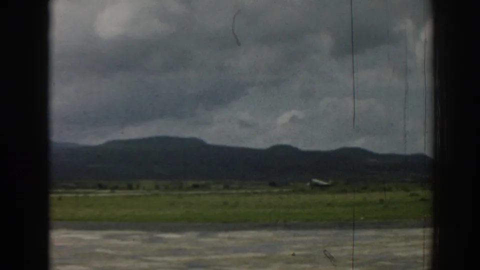 1962 Izmir home-movie frame showing a small propeller airplane near a grassy runway with mountains under gray clouds.