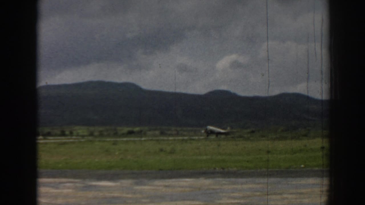 1962 Izmir frame showing a small propeller aircraft low against a grassy airfield and dark mountain horizon.