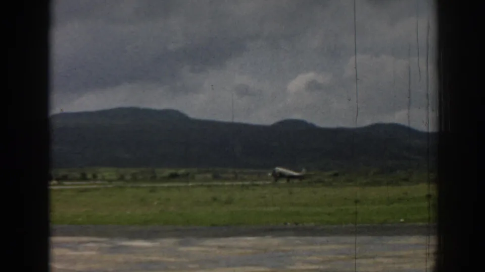 1962 Izmir frame showing a small propeller aircraft low against a grassy airfield and dark mountain horizon.