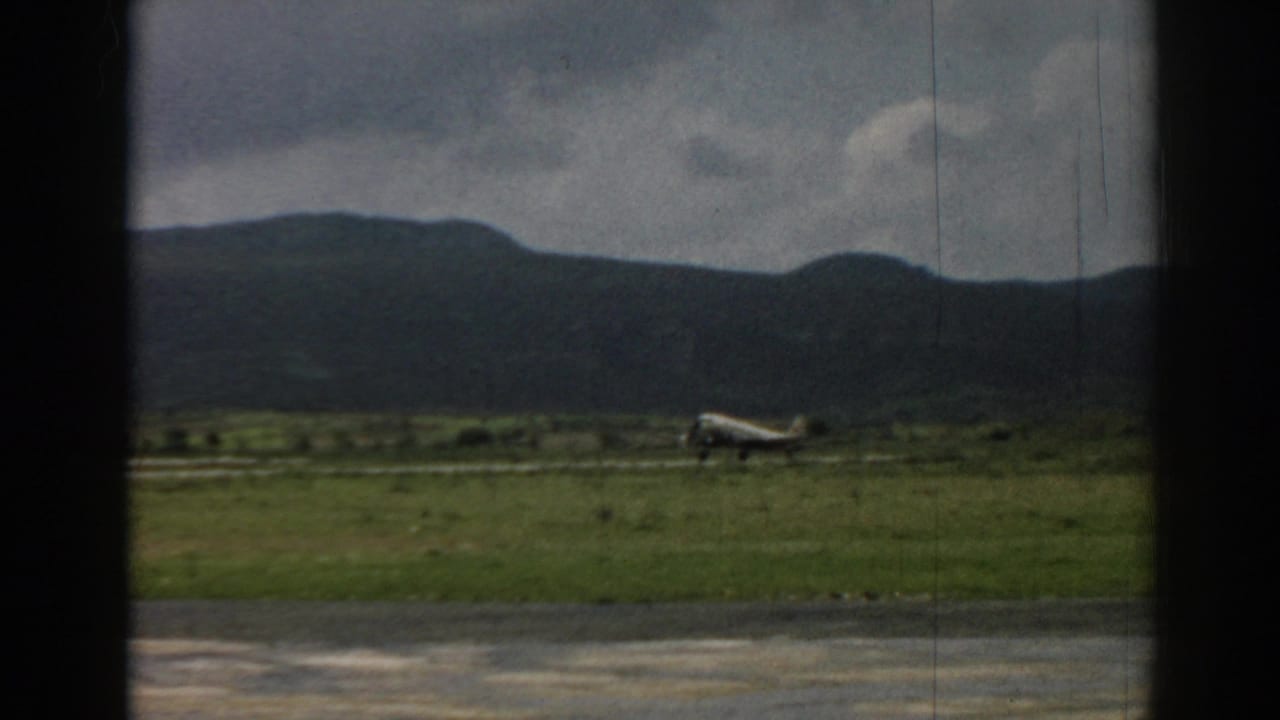 1962 Izmir home-movie frame of a small airplane near takeoff over a grassy airfield with mountains beyond.