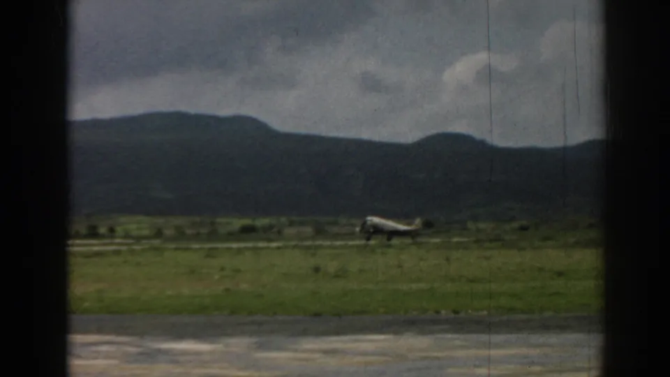 1962 Izmir home-movie frame of a small airplane near takeoff over a grassy airfield with mountains beyond.